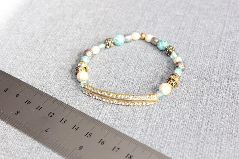 May include: A beaded bracelet with a gold-tone bar embellished with small, clear rhinestones. The bracelet features a mix of white, turquoise, silver, and gold beads. The bracelet is approximately 7 inches in length.