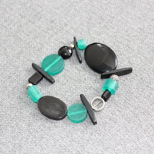 May include: A bracelet featuring a mix of black and teal beads. The bracelet includes oval and round black beads, teal disc-shaped beads, and small black rectangular accents. The bracelet is on a gray surface.