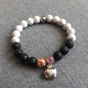 May include: A beaded bracelet featuring white and black lava stone beads. The bracelet includes a silver apple charm, a copper-colored bead, and a pink bead. The beads are strung on an elastic band, creating a stretch bracelet.