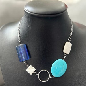 May include: A necklace with a silver chain and a selection of colourful stones. The design incorporates a large, oval turquoise stone, a rectangular blue stone, and white rectangular stones. A silver circle is also included.