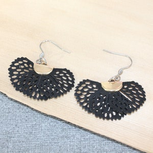 May include: A pair of black, fan-shaped earrings with a delicate, openwork design. The earrings have a gold-colored accent at the top and silver-colored ear wires. The earrings are displayed on a light-colored wooden surface.