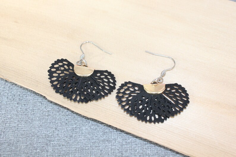 May include: A pair of black, fan-shaped earrings with a delicate, openwork design. The earrings have a gold-colored accent at the top and silver-colored ear wires. The earrings are displayed on a light-colored wooden surface.