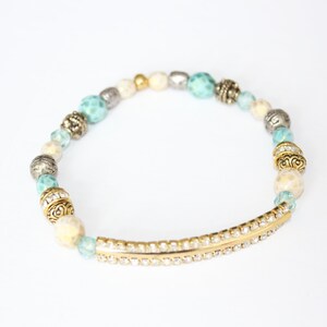 May include: A beaded bracelet featuring a mix of turquoise, cream, silver, and gold beads. The bracelet includes a gold-tone bar embellished with small, clear crystals. The beads are strung on an elastic band.