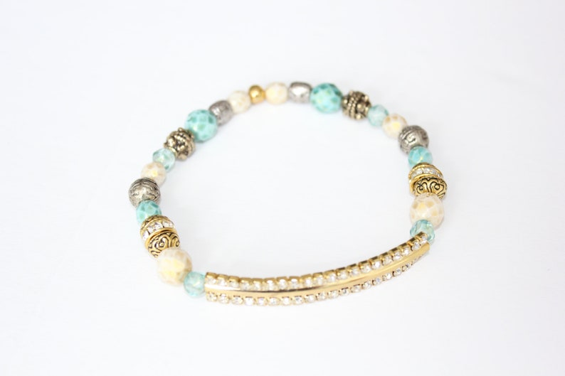 May include: A beaded bracelet featuring a mix of turquoise, cream, silver, and gold beads. The bracelet includes a gold-tone bar embellished with small, clear crystals. The beads are strung on an elastic band.