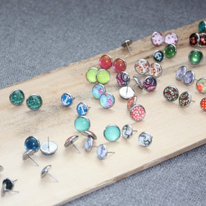 Pu&ograve; includere: Una collezione di orecchini a bottone con vari disegni, tra cui motivi floreali, forme geometriche e colori uniti. Gli orecchini sono esposti su una tavola di legno su sfondo grigio. Gli orecchini hanno perni color argento.