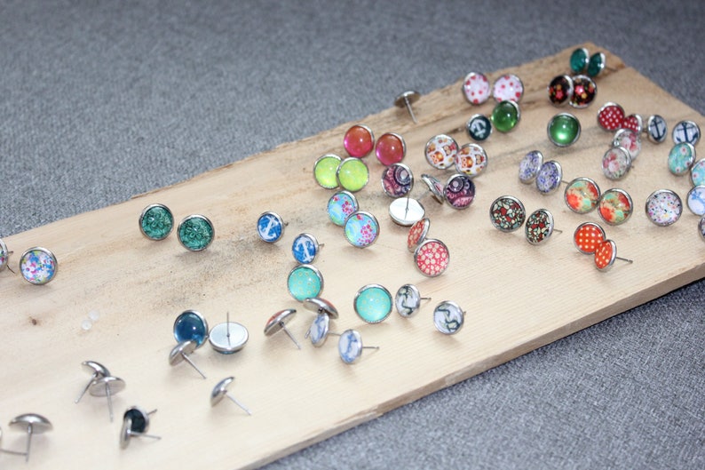 Pu&ograve; includere: Una collezione di orecchini a bottone con vari disegni, tra cui motivi floreali, forme geometriche e colori uniti. Gli orecchini sono esposti su una tavola di legno su sfondo grigio. Gli orecchini hanno perni color argento.