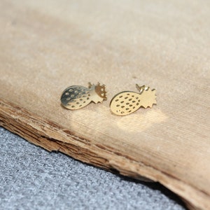 Pu&ograve; includere: Due orecchini a bottone dorati a forma di ananas. Ogni orecchino presenta un corpo strutturato con piccoli dettagli a pois e una corona appuntita. Gli orecchini sono esposti su una superficie marrone chiaro strutturata.