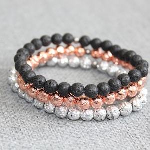 May include: Three beaded bracelets stacked together. The top bracelet features black lava beads, the middle one has rose gold beads, and the bottom one is silver. The bracelets are on a gray surface.