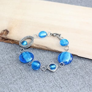 Pu&ograve; includere: Un braccialetto argentato con perline blu rotonde e ovali. Il braccialetto presenta un mix di perline piccole e grandi, con un elemento decorativo ovale e quadrato. Le perline hanno un aspetto strutturato e scintillante.