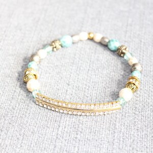 May include: A beaded bracelet featuring a gold-tone curved bar with clear rhinestones. The bracelet includes white, turquoise, and silver beads, along with gold-tone decorative accents. This stretch bracelet is a fashion accessory.