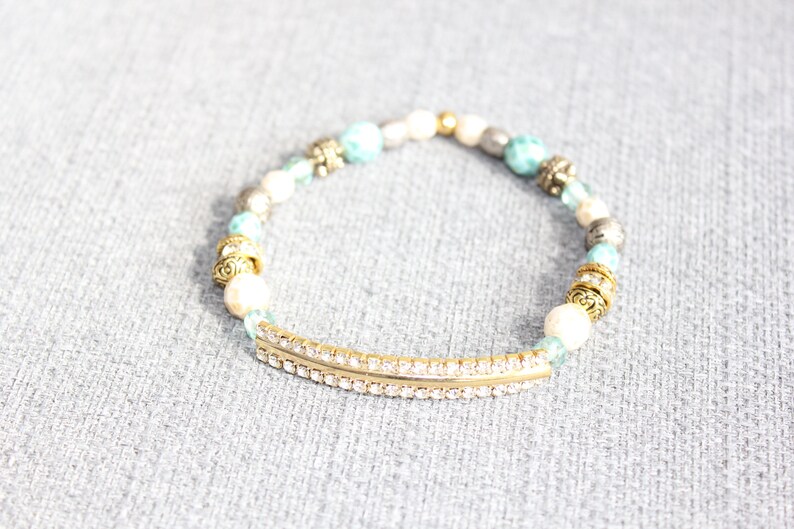 May include: A beaded bracelet featuring a gold-tone curved bar with clear rhinestones. The bracelet includes white, turquoise, and silver beads, along with gold-tone decorative accents. This stretch bracelet is a fashion accessory.