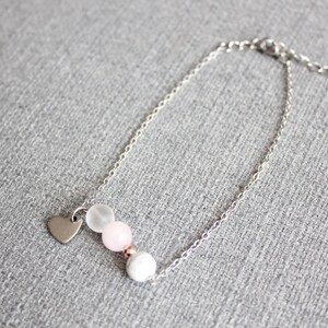 Puede incluir: Una pulsera plateada con un peque&ntilde;o dije de coraz&oacute;n y una serie de cuentas blancas, rosas y transparentes. La pulsera se muestra sobre una superficie gris.