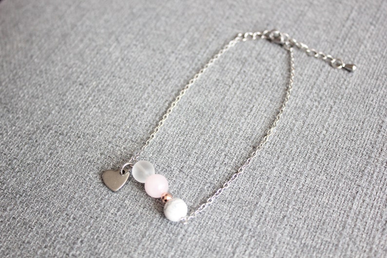 Puede incluir: Una pulsera plateada con un peque&ntilde;o dije de coraz&oacute;n y una serie de cuentas blancas, rosas y transparentes. La pulsera se muestra sobre una superficie gris.