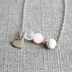 Puede incluir: Un delicado collar de plata con un peque&ntilde;o dije de coraz&oacute;n y una serie de cuentas. Las cuentas incluyen una blanca esmerilada, una rosa p&aacute;lido, una cuenta color cobre y una cuenta blanca con vetas de m&aacute;rmol. El collar se muestra sobre un fondo gris texturizado.