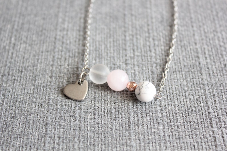 Puede incluir: Un delicado collar de plata con un peque&ntilde;o dije de coraz&oacute;n y una serie de cuentas. Las cuentas incluyen una blanca esmerilada, una rosa p&aacute;lido, una cuenta color cobre y una cuenta blanca con vetas de m&aacute;rmol. El collar se muestra sobre un fondo gris texturizado.