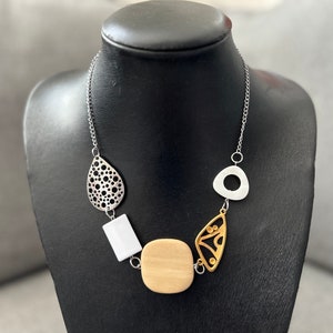 May include: A silver necklace with assorted geometric pendants. The necklace includes a teardrop-shaped pendant with a perforated design, a white rectangular pendant, a light wood square pendant, a gold-toned leaf-shaped pendant, and a silver triangle pendant.