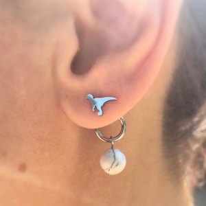 May include: A light blue dinosaur stud earring and a silver hoop earring with a white and gray marbled bead. The earrings are worn on the ear. The dinosaur stud is a small, stylized silhouette.