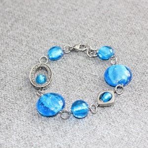 Pu&ograve; includere: Un braccialetto argentato con una serie di perline rotonde di vetro blu. Il braccialetto ha una chiusura a moschettone e include un elemento ovale decorativo con una perlina blu pi&ugrave; piccola. Le perline sono di un blu vibrante.
