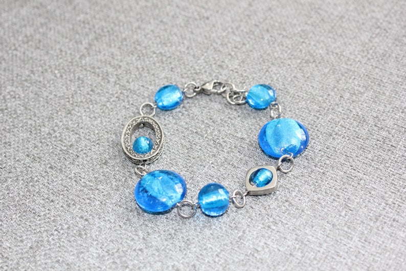 Pu&ograve; includere: Un braccialetto argentato con una serie di perline rotonde di vetro blu. Il braccialetto ha una chiusura a moschettone e include un elemento ovale decorativo con una perlina blu pi&ugrave; piccola. Le perline sono di un blu vibrante.