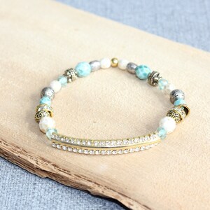 May include: A beaded bracelet featuring a gold-tone bar with clear rhinestones, interspersed with turquoise, white, and silver beads. The bracelet has decorative gold-tone accents and is displayed on a light brown wooden surface.