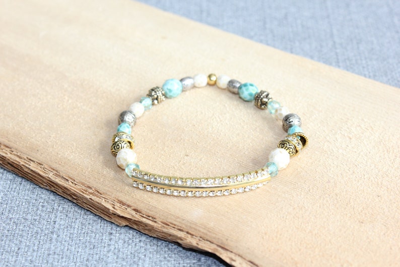 May include: A beaded bracelet featuring a gold-tone bar with clear rhinestones, interspersed with turquoise, white, and silver beads. The bracelet has decorative gold-tone accents and is displayed on a light brown wooden surface.