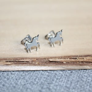 May include: A pair of silver-colored horse-shaped stud earrings. The earrings are small, with a simple outline of a horse. They are displayed on a light brown wooden surface, with a gray fabric in the background.