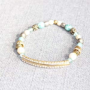 May include: A beaded bracelet featuring a gold-tone bar with clear rhinestones, interspersed with white, turquoise, silver, and gold beads. The bracelet has decorative gold-tone accents and is set against a gray background.