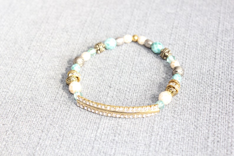 May include: A beaded bracelet featuring a gold-tone bar with clear rhinestones, interspersed with white, turquoise, silver, and gold beads. The bracelet has decorative gold-tone accents and is set against a gray background.