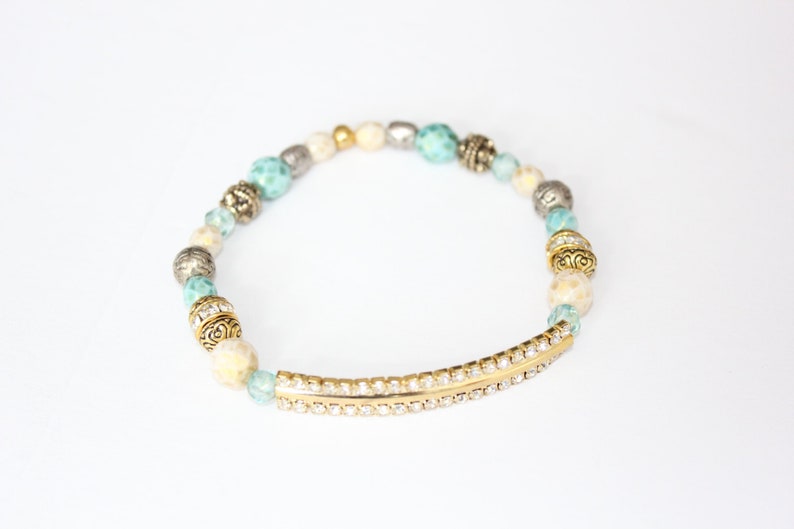 May include: An elegant bracelet featuring a mix of beads in cream, light blue, silver, and gold. The bracelet includes faceted beads, ornate gold-toned beads, and a gold-toned bar embellished with small, clear crystals. The bracelet is designed to be worn on the wrist.