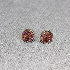 Puede incluir: Un par de pendientes de botón redondos con un engaste de metal plateado. Los pendientes presentan una superficie brillante y texturizada con tonos de rosa, naranja y dorado. Los pendientes se muestran sobre una superficie gris texturizada.
