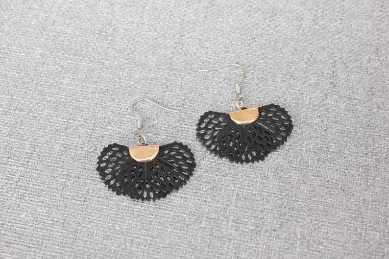 May include: A pair of black, fan-shaped earrings with a gold-colored accent at the top. The earrings have a delicate, openwork design and silver-colored ear wires. The earrings are displayed on a gray textured surface.