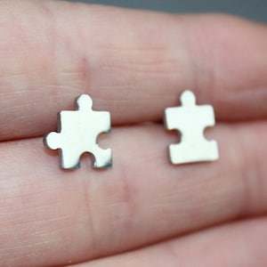May include: A pair of silver-colored puzzle piece stud earrings. The earrings are small, with a simple, minimalist design. The earrings are shown on a person's hand.