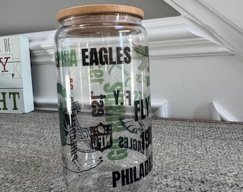 May include: A clear glass jar with a bamboo lid. The jar is decorated with a Philadelphia Eagles theme, featuring the team's logo, the words "Fly Eagles Fly", and the team's name.