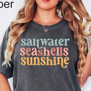 May include: Dark gray t-shirt with the words "saltwater seashells sunshine" in teal, coral, and yellow. The shirt is a relaxed fit and has short sleeves. The word "Pepper" is in the top left corner.