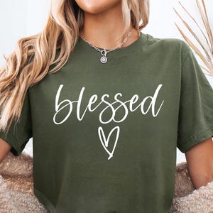 May include: Olive green t-shirt with the word "blessed" in white script and a white heart. The shirt is part of a casual outfit, paired with denim shorts and a cream-colored cardigan. The shirt is labeled "Military Green".