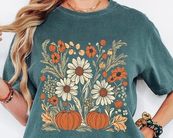 May include: Blue Spruce t-shirt featuring a floral and pumpkin design. The graphic includes white daisies, orange flowers, and two pumpkins. The text "Blue Spruce" is printed at the top. The shirt is a relaxed fit.