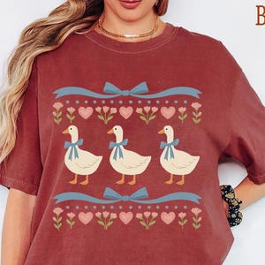 May include: Brick-colored t-shirt featuring a whimsical design of three white geese adorned with blue bows. The geese are centered on the shirt, above a row of pink hearts and flowers. The word "BRICK" is printed in brown at the top right.