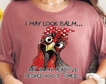 May include: Heather mauve t-shirt with a humorous graphic of a chicken wearing a red bandana and glasses. The text reads, "I MAY LOOK CALM... BUT IN MY HEAD I'VE PECKED YOU 3 TIMES..." A fun, casual tee.