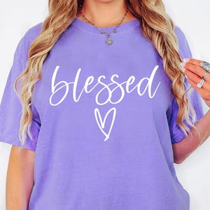 May include: A purple oversized t-shirt with the word "blessed" and a heart printed in white on the front.