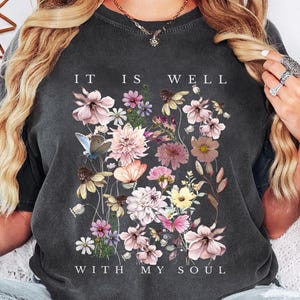 May include: A charcoal grey short-sleeve t-shirt featuring a graphic print of various colourful flowers, butterflies, and the text "IT IS WELL WITH MY SOUL" in white lettering. The shirt has a crew neckline and a faded, vintage-inspired fabric finish.