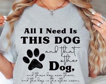 May include: Light gray t-shirt with the text "All I Need Is THIS DOG and that other Dog." A black paw print graphic is below the text. The shirt is a crew neck style.
