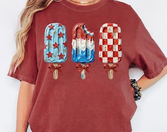 Peut inclure: Un t-shirt couleur brique orné d'un motif représentant trois sucettes glacées, chacune nouée d'un petit ruban rouge. La sucette de gauche est bleue avec des étoiles rouges, celle du centre est rouge, blanche et bleue, et celle de droite est à damier rouge et blanc.