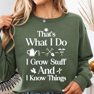 May include: A green sweatshirt with the text "That's What I Do I Grow Stuff And I Know Things" in white. The text is surrounded by gardening icons, including a watering can, a shovel, and a plant.