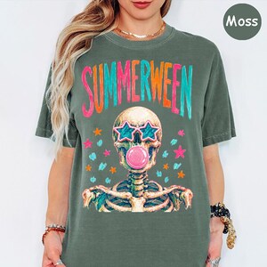 May include: A moss green t-shirt featuring a graphic of a skeleton wearing star-shaped sunglasses and blowing a pink bubblegum bubble. The text "SUMMERWEEN" is printed in colorful letters above the skeleton, surrounded by small star icons.