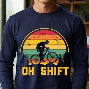 May include: Navy blue long-sleeve shirt with a graphic of a cyclist riding a bicycle, set against a sunset design with the text "OH SHIFT" in yellow. The design includes trees and birds.