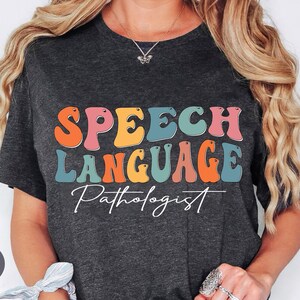 May include: A heather dark gray t-shirt featuring the text SPEECH LANGUAGE in a colorful, wavy, retro-style font, with the word Pathologist written below in a white cursive script. The shirt is short-sleeved with a crew neckline.