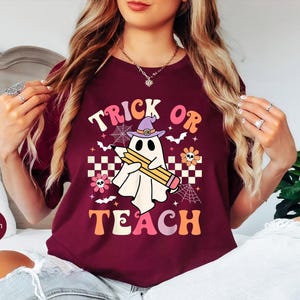 May include: Maroon t-shirt with a Halloween-themed graphic. The design features a ghost wearing a witch hat and holding a pencil, with the words "Trick or Teach" in a retro font. Additional elements include bats, flowers, and spiderwebs.