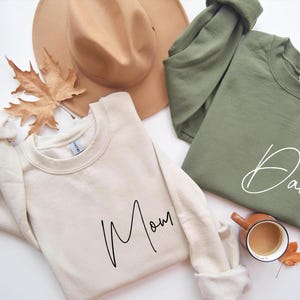 May include: Two cream colored sweatshirts with black script. One says "Mom" and the other says "Dad".
