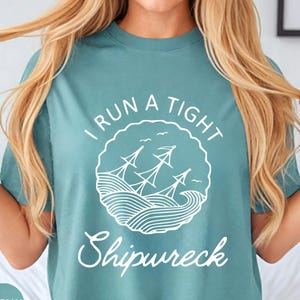 May include: Seafoam green t-shirt with the text "I RUN A TIGHT Shipwreck" in white. The graphic features a ship on wavy water inside a circular frame. The shirt is a solid color and has a crew neck.