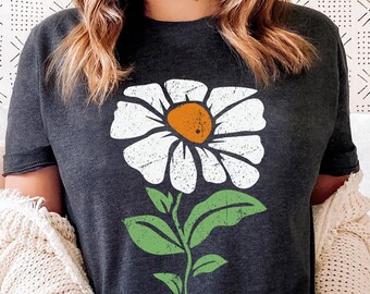 May include: A dark grey heather t-shirt with a white daisy graphic. The daisy has a yellow center and green leaves. The shirt is a relaxed fit and has a crew neck.